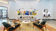 Located on Bridgeport Way in Suffolk, Virginia, this single-floor facility is a thoughtfully designed communal space that blends contemporary aesthetics with functional versatility. Polished concrete floors extend throughout much of the space, contrasted by rich hardwood deck-style flooring in the main lounge area, lending warmth and visual interest to the open plan layout.

The main gathering area is expansive and bright, anchored by a fireplace and wall-mounted television, with a curated seating arrangement featuring neutral sofas, mid-century modern accent chairs, and a vibrant geometric area rug. Bold, colorful abstract artwork adorns the white paneled walls, adding an artistic focal point. A bar-height counter with stools and pendant lighting creates a casual social hub nearby, while a foosball table underscores the recreational nature of the space.

Floor-to-ceiling steel-framed glass partitions and oversized windows flood the interior with natural light and offer views of mature trees outside. A glass garage-style door opens the space to an outdoor area, enhancing indoor-outdoor flow. A dedicated conference room features a large display screen and a sleek oval table. A cozy lounge nook with paired armchairs and a coffee table provides a quieter setting. Lush snake plants line interior corridors, contributing a fresh, welcoming atmosphere throughout.