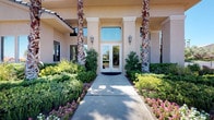 This single-story building makes a striking first impression with a beautifully landscaped entrance walkway lined with lush shrubs, colorful flowering plants, and towering palm trees. Double glass doors framed by classical columns and decorative wall sconces invite visitors inside, where the architecture immediately commands attention.

The interior centers around a dramatic rotunda lobby distinguished by a tiered, coffered ceiling with recessed lighting and warm ambient uplighting that highlights the curved architectural details above. The space is finished with large-format marble-look tile flooring, lending an air of elegance and openness. Generous windows on multiple walls flood the room with abundant natural light and offer views of the surrounding greenery, creating a bright and airy atmosphere.

The lobby is thoughtfully furnished with dark leather sofas and armchairs accented by colorful throw pillows, arranged around a sleek glass-and-metal coffee table. A built-in niche along the back wall serves as a focal display area, showcasing decorative artwork and cabinetry. Tall potted plants positioned throughout the space add a natural, welcoming touch. Wall-mounted decorative sconces complement the overall refined aesthetic, making this a polished and functional gathering space that balances sophistication with comfort.