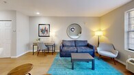 Located at 8710 Research Dr in Charlotte, NC, this efficient one-bedroom, one-bathroom unit offers a thoughtfully arranged single-floor layout that maximizes every square foot. The main living and sleeping area is the heart of the space, combining a full-sized bed, a comfortable seating area with a sofa, armchair, and coffee table, plus a dedicated workspace with a desk and chair — all unified by light wood-look flooring and a vibrant teal area rug that adds a pop of color. A large window with slatted blinds allows natural light to brighten the room, while wall-mounted art and a round mirror contribute to a clean, modern aesthetic. A wall-unit HVAC system provides individual climate control. The compact kitchen is efficiently designed with white shaker-style cabinetry, white subway tile backsplash, light stone countertops, a stainless steel sink, a dishwasher, and a full-sized refrigerator — offering essential cooking and storage functionality. The bathroom features a clean, bright design with a full soaking tub and shower combo, a white vanity with under-sink storage, and warm wood-look flooring. The entrance provides a natural transition into the living space, completing this well-organized, functional unit.