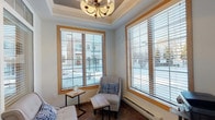 This single-floor space is thoughtfully designed as a comfortable common area, combining a bright seating lounge with a practical WiFi room. The main lounge area showcases an elegant tray ceiling accented by a stylish chandelier, creating a refined architectural focal point overhead. Large windows framed in warm natural wood trim flood the space with natural light and offer views of the surrounding exterior, while horizontal blinds provide adjustable privacy. The flooring throughout is laid in durable stone-look tile, lending a clean and cohesive look. The seating nook is furnished with cushioned chairs and compact side tables, creating an inviting spot for relaxation or informal conversation. A dedicated work station with a printer and supply organizer makes the space functional for residents or guests needing everyday office amenities. A glass-paneled interior door leads to the WiFi room, which features cabinetry with granite-style countertops and additional workspace. The building entry area includes a secure glass door with transom windows, fire alarm and access control panels, and warm oak-toned doors, reflecting a well-maintained and safety-conscious environment. Baseboard heating units ensure year-round comfort throughout the space.