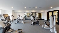 This fitness center offers a thoughtfully organized single-level layout designed to support a comprehensive workout experience. The space is divided into distinct functional zones, each outfitted with professional-grade equipment. The cardio area is positioned along a wall of large, floor-to-ceiling windows that flood the space with natural light and provide open views of the surrounding exterior, creating an energizing atmosphere for treadmill, elliptical, and stationary bike workouts. Matrix-brand treadmills and ellipticals with individual screens are prominently featured in this section.

The strength training area occupies a generous portion of the facility and is lined with full-length mirrors that enhance the sense of depth and allow users to monitor their form. A variety of Hoist and Matrix selectorized cable and resistance machines are arranged throughout this zone, providing options for a full-body workout. Recessed lighting and white walls contribute to a clean, modern aesthetic throughout.

Toward the rear of the space, a locker room area is accessible, complete with individual lockers and water fountain facilities. The flooring throughout consists of durable, textured grey carpet tile suited for high-traffic fitness use. The overall design is functional, modern, and well-lit, fostering a professional gym environment.