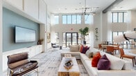 This single-floor community clubhouse is designed with a contemporary aesthetic, combining open-concept layouts with thoughtful architectural details throughout. The main lounge area is anchored by dramatic double-height ceilings and an expansive wall of black-framed windows that flood the space with natural light while offering direct views of the outdoor pool area beyond. A large sectional sofa in neutral tones, accented with bold jewel-toned throw pillows, anchors the central seating area around a live-edge wood coffee table, while a statement geometric chandelier overhead adds a striking visual focal point. A teal accent wall frames a large wall-mounted television set above custom white cabinetry, flanked by a built-in shelving unit displaying curated decorative objects. Rich leather lounge chairs add warmth and contrast to the light-toned carpet throughout the lounge. An adjacent kitchen and bar area features a herringbone-patterned hardwood floor, soaring ceilings with exposed dark beams, pendant lighting, and a generous island with bar seating — ideal for social gatherings. A glass-partitioned flex room with hardwood flooring offers additional versatile space. Together, these areas create a welcoming, upscale environment that balances comfort, style, and functionality.