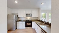 Located at 150 Graylawn Ave in Petaluma, California, this 2-bedroom, 1-bathroom home offers a thoughtfully designed single-floor layout with a bright and clean aesthetic throughout. The kitchen is a standout feature, showcasing crisp white shaker-style cabinetry extending to the ceiling, dark countertops, stainless steel appliances including a refrigerator, electric range, and over-the-range microwave, and a stainless steel sink positioned beneath a window that brings in natural light. Light wood-style flooring runs through the main living areas, complementing the home's modern, neutral palette. The open living room is airy and well-lit, featuring a ceiling fan and a wall-mounted heating unit. The spacious primary bedroom offers generous proportions with soft carpet flooring, a mirrored sliding closet, and built-in shelving. The second bedroom features carpet flooring and a sliding glass door that opens directly onto the private balcony, which overlooks mature trees and an open field — a serene outdoor retreat. The bathroom is efficiently laid out with a tub/shower combination, a vanity with a countertop sink, and wood-style flooring. Two hallways connect the spaces smoothly, and the overall ambiance is clean, neutral, and move-in ready.