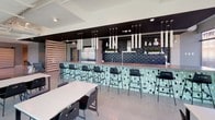 Wheelhouse is a thoughtfully designed co-living community amenity floor that seamlessly blends social, work, and leisure environments into a single, open-plan space. Large floor-to-ceiling windows line the street-facing facade, flooding the interior with abundant natural light and offering dynamic views of the surrounding urban streetscape, including a visible church steeple and active city streets below.

The lounge area anchors the social heart of the space, featuring comfortable seating arrangements with upholstered armchairs, a plush sofa, and patterned area rugs atop polished concrete floors. Exposed ductwork and high ceilings contribute to a contemporary industrial aesthetic throughout. Sliding barn doors provide flexible room separation, allowing the open layout to be configured for various uses.

A dedicated co-working zone offers multiple tables and chairs positioned against the expansive branded windows, creating a bright and productive work environment. Adjacent to this is a striking communal kitchen and bar area, highlighted by a long counter with a decorative diamond-patterned tile facade in mint and navy tones, a dark tiled backsplash, and a dramatic array of pendant lighting hanging from above. The stainless steel appliances and wood-accented shelving complete the polished, design-forward aesthetic that defines this vibrant community hub.