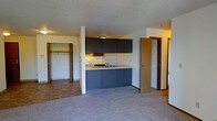 This single-floor space offers an open, functional layout that seamlessly connects the kitchen and living areas. The kitchen is outfitted with dark navy ribbed cabinetry, both upper and lower, complemented by a light-toned countertop and a built-in sink. A matching breakfast bar extends outward, providing a casual dining or workspace surface. Under-cabinet lighting adds a warm glow to the cooking area, while overhead recessed lighting illuminates the broader living space. The textured ceiling adds a classic architectural detail throughout. Plush carpeting covers the living area, lending a comfortable and warm ambiance underfoot. A generously sized closet with an open doorway reveals ample hanging and shelving storage, ideal for organization. An additional door on the opposite side of the kitchen hints at further rooms or utility spaces beyond. Warm wood-toned door and trim accents provide a cohesive, grounded aesthetic throughout the unit. The overall design is practical and well-organized, making efficient use of the available floor plan while maintaining a comfortable, livable atmosphere.