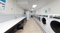 This single-floor laundromat is designed with functionality and user comfort in mind. The space features a long row of commercial-grade front-load washing machines and dryers lining one wall, offering ample capacity to handle multiple loads simultaneously. The machines are clearly numbered for easy identification, and instructional signage is posted on the walls to guide users through the laundry process.

Opposite the machines, extended folding tables provide generous workspace for sorting and folding laundry, accompanied by rolling laundry carts for added convenience. The clean, light-toned walls are accented with a two-tone wainscoting detail, contributing to a tidy and welcoming atmosphere. A large interior window connects to an adjacent room, enhancing visibility and openness throughout the space.

The flooring consists of light wood-look planks, giving the facility a clean and modern aesthetic. Overhead, recessed panel lighting and skylight-style flush fixtures ensure the space is evenly and brightly lit throughout. A wall clock keeps users informed of the time, and an informational bulletin board provides additional guidance. The overall layout is spacious and thoughtfully organized, making it easy to navigate and comfortable for extended visits.