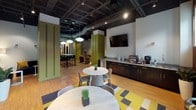 This single-floor workspace is thoughtfully designed to accommodate a variety of work and social activities within one cohesive environment. The interior adopts an industrial-chic aesthetic, with exposed black ceilings revealing ductwork and structural elements overhead, contrasted by warm bamboo hardwood flooring that runs throughout the space.

The layout is divided into several functional zones. A central café-style area features round marble-top tables paired with modern chairs, set atop a graphic yellow, grey, and black area rug. A built-in credenza along one wall houses a coffee station and small appliances, making it a natural gathering point. Adjacent to this, a tall communal table with black bar stools sits beneath a pair of iconic globe pendant lights, suited for collaborative work or casual meetings.

Olive-green paneled columns and partition walls define separate lounge areas furnished with mustard and navy upholstered seating. A dedicated nook features floor-to-ceiling wood slat wall paneling, natural window light, curated artwork, and an intimate round table — ideal for focused conversations. Wall-mounted flat-screen TVs are positioned throughout, supporting both presentation and entertainment needs. The overall ambiance is polished yet inviting.