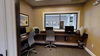 This business center is a thoughtfully designed, single-floor workspace built to accommodate productivity and convenience. The room features a continuous built-in wooden desk along the walls, providing ample workspace for multiple users simultaneously. Three computer workstations are set up along the desk, each equipped with monitors, keyboards, and accompanying peripherals, ensuring guests have reliable access to technology. A printer and additional office equipment are also available, making the space fully functional for a range of business needs.

The interior is finished with warm, neutral-toned walls that create a calm and professional atmosphere, complemented by a drop ceiling with recessed lighting that ensures consistent, comfortable illumination throughout. Carpeted flooring in a muted tone adds a layer of quiet and comfort underfoot. Ergonomic task chairs at each workstation prioritize user comfort during extended use.

A large interior sliding window serves as a standout architectural feature, offering a clear view into the adjacent common area, which is decorated with framed artwork and finished in a light, welcoming palette. Framed wall art within the business center itself adds a polished, hospitality-oriented touch, making this space as visually inviting as it is practical.