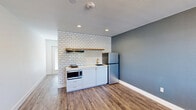 Located at 645 W Pullman Rd in Moscow, Idaho, this efficiently designed studio unit offers a thoughtful layout across a single floor, comprising a combined living area, a hallway, and a full bathroom. The living space features warm wood-tone laminate flooring and clean white walls, creating a bright, neutral backdrop that maximizes the sense of openness. A well-appointed kitchenette lines one wall, showcasing white subway tile backsplash, white cabinetry with gold-tone hardware, a countertop range, microwave, stainless steel refrigerator, and a sink — all accented by a floating wood shelf above. Recessed lighting and a slate-blue accent wall add depth and a modern touch to the kitchen area. The bathroom is a standout feature, finished with large-format white wall tiles, dark grout, hexagonal mosaic floor tiles, and a matte black shower fixture, delivering a contemporary, design-forward aesthetic. A private balcony accessible from the unit extends the living space outdoors, offering composite decking, metal railing, and open views of surrounding greenery and the neighborhood skyline. This unit combines efficient use of space with modern finishes throughout.
