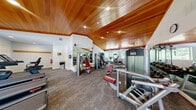 Located at 7 Kimball Ct in Woburn, Massachusetts, this fitness center offers a thoughtfully designed exercise space within a single open floor plan. A defining architectural feature is the striking vaulted ceiling clad in warm-toned wood paneling, which lends the room a refined, inviting character uncommon in typical gym settings. Recessed lighting throughout complements the natural light streaming in from multiple large windows, which frame views of surrounding greenery and create a bright, energizing atmosphere.

The exercise room is comprehensively outfitted with a diverse array of equipment to support a full-body workout routine. The cardio section features multiple Star Trac treadmills, elliptical trainers, and a stationary bike, all positioned near the windows to take advantage of the natural light and outdoor views. The strength training area includes a variety of selectorized resistance machines with red-accented seats, a cable cross machine, adjustable benches, and a well-stocked dumbbell rack ranging across multiple weight increments.

Full-length mirrors line sections of the walls, enhancing the sense of spaciousness and allowing users to monitor their form during workouts. The floor is finished with durable, commercial-grade carpet. Safety features including a fire extinguisher and clearly marked exit signage are also present throughout the space.