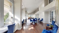 Located at 601 Rosery Rd NE in Largo, Florida, this single-floor community amenity space offers a well-appointed and multi-functional layout designed for both socializing and relaxation. Rich hardwood flooring runs throughout, anchoring the space with warmth and elegance.

Soaring double-height ceilings create a dramatic sense of openness, while large windows along the perimeter flood the interior with abundant natural light and offer pleasant views of the surrounding Florida landscape, including swaying palms and lush greenery.

The dining area serves as a central gathering point, featuring a sleek white rectangular table surrounded by plush, tufted royal blue velvet chairs — a bold design choice that lends a vibrant, upscale character to the room. Artwork on the walls adds a tasteful finishing touch to this inviting area.

Adjacent to the dining space, a recreation zone accommodates a full-size billiards table set atop gleaming hardwood floors, with a wall-mounted television nearby for entertainment. French doors with glass panels provide easy access to the outdoors and further enhance the bright, airy atmosphere.

A quiet seating alcove lined with tall windows and neutral accent chairs offers a comfortable retreat, ideal for reading or casual conversation. An internet café area with individual workstations rounds out this thoughtfully designed, community-focused space.
