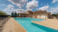 Located at 9123 Cleveland St in Merrillville, Indiana, this outdoor amenity space centers around a generously sized swimming pool with a distinctive curved design and wide, shallow entry steps for easy access. The pool's crystal-clear blue water is complemented by an expansive concrete sundeck that provides ample space for lounging, lined with sling-back lounge chairs and umbrellas for shade.

A charming pergola structure anchors one end of the deck, offering a covered gathering space with additional seating ideal for relaxation or socializing. On the opposite side, a well-appointed pool house with a pitched roof, white garage-style storage doors, and classic column detailing adds architectural character to the setting. Ornamental brick pillars mark the entrance area, enhancing the overall aesthetic.

The perimeter is enclosed by black iron fencing, offering both security and an unobstructed view of the surrounding residential buildings and mature trees. Colorful plantings and garden beds add natural beauty along the edges of the space. The open layout and abundance of seating arrangements make this a versatile outdoor destination, well-suited for both quiet relaxation and community gatherings under the open Indiana sky.
