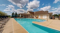 Located at 9123 Cleveland St in Merrillville, Indiana, this outdoor amenity space centers around a generously sized swimming pool with a distinctive curved design and wide, shallow entry steps for easy access. The pool's crystal-clear blue water is complemented by an expansive concrete sundeck that provides ample space for lounging, lined with sling-back lounge chairs and umbrellas for shade.

A charming pergola structure anchors one end of the deck, offering a covered gathering space with additional seating ideal for relaxation or socializing. On the opposite side, a well-appointed pool house with a pitched roof, white garage-style storage doors, and classic column detailing adds architectural character to the setting. Ornamental brick pillars mark the entrance area, enhancing the overall aesthetic.

The perimeter is enclosed by black iron fencing, offering both security and an unobstructed view of the surrounding residential buildings and mature trees. Colorful plantings and garden beds add natural beauty along the edges of the space. The open layout and abundance of seating arrangements make this a versatile outdoor destination, well-suited for both quiet relaxation and community gatherings under the open Indiana sky.