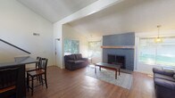This single-floor space offers an open, well-connected layout defined by notable architectural character and warm, inviting finishes throughout. The living area serves as the heart of the space, showcasing vaulted ceilings with exposed beam detailing and a textured ceiling that adds visual depth. A bold, floor-to-ceiling gray brick fireplace with a solid wood mantel acts as the room's centerpiece, flanked by multiple windows fitted with blinds that allow ample natural light to filter in while framing views of the surrounding greenery outdoors. Rich, dark hardwood flooring runs continuously throughout, lending warmth and cohesion to the open floor plan. The living room flows naturally into a dining area, where pendant lighting hangs above dining tables, creating a distinct yet connected gathering space. A bar-height counter with seating provides an additional dining option. Vaulted ceilings carry through to portions of the dining and living zones, contributing to an overall sense of spaciousness and volume. Track lighting in the dining area adds functional illumination, while the open sightlines between zones enhance the communal feel of the space. Overall, the combination of architectural detailing, natural light, and hardwood floors creates a comfortable and well-appointed environment suited for both relaxing and entertaining.