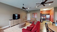 This single-floor space offers a thoughtfully designed open-concept layout that connects the living area and kitchen in a functional and inviting arrangement. The living area is generously sized, anchored by warm hardwood flooring and a large area rug that defines the seating zone. Crown molding runs along the ceiling, adding a refined architectural touch, while two ceiling fans ensure comfortable airflow throughout the space. Recessed lighting complements the overall illumination, contributing to a bright and welcoming ambiance. The living room features ample seating, a spacious media console, and coordinating accent pieces that create a cohesive, relaxed aesthetic. A vibrant piece of wall art adds a pop of color against the neutral taupe walls. Adjacent to the living area, the kitchen is visible and features dark cabinetry, modern appliances, and a bold accent wall in a warm orange hue, providing a striking contrast to the more subdued tones of the main living space. A private door off the living area leads to additional spaces, reinforcing the practical flow of the floor plan. Overall, the space combines comfort and functionality with tasteful design details throughout.