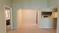This single-floor space features an open and functional layout designed to maximize usability. The main living area is finished with wall-to-wall carpeting in a neutral beige tone, creating a warm and understated foundation that complements the light-colored walls throughout. A wide arched passageway connects the primary living space to an adjacent room, which benefits from recessed ceiling lighting that adds a focused, ambient glow. White baseboards run consistently along the walls, contributing to a clean and cohesive aesthetic. To the right, a pass-through or open counter provides a direct sightline into the kitchen, which features dark wood cabinetry, a black refrigerator, and a range with an overhead microwave or vent hood. The kitchen's light countertops contrast neatly with the darker appliances and cabinetry. The overall color palette of soft greens and warm neutrals gives the space a calm, move-in-ready feel. The layout promotes easy flow between the living and kitchen areas, making it well-suited for everyday living.