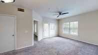 This single-floor residential unit offers a well-organized layout designed for comfortable everyday living. The spacious living area is finished with plush carpeting and features a ceiling fan, multiple windows with horizontal blinds that invite an abundance of natural light, and a glass-paneled exterior door providing direct outdoor access to a private balcony or patio. The open-plan kitchen is equipped with white appliances including a refrigerator, electric range, and built-in microwave, complemented by warm wood-toned cabinetry, light-colored countertops, a double sink, and track lighting overhead. A breakfast bar connects the kitchen to the adjoining living space, making it ideal for casual dining or entertaining. The bedrooms are generously sized, each carpeted and fitted with ceiling fans and windows that offer garden views. One bedroom connects directly to a private bathroom, which features a single-sink vanity with a light-toned countertop, wood-look flooring, a full tub-shower combination with tile surround, and a large mirror. Additional rooms provide flexible space for various uses. Throughout the unit, neutral-toned walls and clean white trim create a bright, cohesive aesthetic.