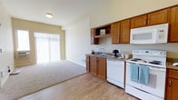 This single-floor unit offers an open layout that seamlessly connects the kitchen and living area, creating a functional and welcoming atmosphere. The kitchen is outfitted with warm honey-toned wood cabinetry, providing ample upper and lower storage. White appliances, including a range, built-in microwave, and double-basin sink, line the kitchen wall, complemented by a laminate countertop and easy-to-maintain hardwood-style flooring. The transition from the kitchen into the living space is marked by a shift to plush carpeting, adding comfort underfoot in the lounge area. A large sliding glass door fitted with vertical blinds anchors the far wall, allowing generous natural light to pour in and offering access to an outdoor space. The neutral, light beige wall palette throughout enhances the brightness of the unit and provides a versatile backdrop for various décor styles. The layout is efficiently designed, making the most of the available space while maintaining an open, airy feel suitable for everyday living.