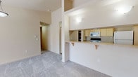 This single-floor residence offers a well-organized layout designed for comfortable everyday living. The open-concept living area is generously proportioned, featuring plush wall-to-wall carpeting, vaulted ceilings, and a ceiling fan with pendant lighting. A sliding glass door and a large window flood the space with natural light and provide views of the surrounding greenery outside.

The kitchen is positioned adjacent to the living area and is defined by a raised breakfast bar with decorative wood corbels, providing both a casual dining option and a natural boundary between spaces. Light wood cabinetry, laminate countertops, tile flooring, and a full suite of appliances — including a refrigerator, electric range, microwave, and dishwasher — make the kitchen fully functional. A dedicated laundry room is conveniently accessible directly from the kitchen and houses a full-size washer and dryer.

The two bedrooms are carpeted and well-lit by large windows, each offering a closet with built-in shelving for ample storage. The bathroom is finished with tile flooring, a light wood vanity, and a tub-shower combination enclosed by sliding frosted glass doors, along with a grab bar for added accessibility. Neutral warm tones throughout the space create a calm and cohesive aesthetic.