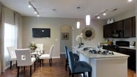 This single-floor residence showcases a thoughtfully designed open-concept layout that seamlessly connects the kitchen, dining, and living areas. The kitchen is a standout feature, boasting a spacious granite-topped island with built-in sink and seating for multiple bar-stool chairs upholstered in teal, creating a vibrant focal point. Pendant lighting hangs above the island, complementing the track lighting that runs throughout the space and ensures ample illumination. Dark espresso cabinetry provides rich contrast against the light granite countertops and neutral gray-toned walls. The adjacent dining area features a glass-top table surrounded by white upholstered chairs, adding a touch of elegance. A wall-mounted flat-screen television and a decorative sunburst mirror with floral accents contribute to the refined ambiance of the living and dining zones. Rich, dark hardwood flooring flows throughout, grounding the space with warmth and continuity. An arched interior passageway adds an architectural detail that lends character to the overall design. The cohesive color palette of neutral walls, dark wood tones, and bold teal accents creates a modern yet inviting atmosphere throughout this well-appointed residence.