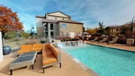 This community amenity space offers residents two distinct areas for recreation and wellness. The outdoor pool area is a standout feature, showcasing a sparkling, generously sized swimming pool with decorative fountain jets along its edge. The surrounding pool deck is furnished with comfortable lounge chairs and wicker seating with vibrant orange cushions, creating an inviting atmosphere for relaxation. Mature landscaping and ornamental plantings frame the space, while the backdrop of a handsome stone-and-siding building adds architectural character. Umbrellas and additional seating areas provide shaded options throughout the deck.

The adjacent fitness center is a well-organized, single-level space designed to support a comprehensive workout routine. Bold orange and blue accent stripes run along the upper walls, lending the room an energetic ambiance. Large windows overlook the pool area, filling the space with natural light. The fitness center is outfitted with a full array of cardio equipment, including treadmills, upright and recumbent bikes, and an elliptical trainer, as well as a multi-station strength machine, a weight bench, and a rack of free weights supported by a wall-mounted mirror. Patterned carpet tile flooring, ceiling fans, and recessed lighting complete the space, ensuring a comfortable and functional environment for residents.