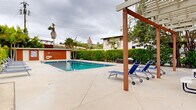 Located at 3902-3920 Lenawee Ave in Culver City, California, this outdoor amenity space centers around a generously sized swimming pool with clear, blue water and a classic tiled border. The pool deck is finished in light-toned concrete, providing ample space for relaxation and movement throughout the area. A row of blue lounge chairs lines one side of the pool, offering residents a comfortable place to unwind, while a wooden pergola structure at one end adds architectural interest and a shaded gathering spot. A prominent red-sided utility building sits at one end of the pool area, housing what appear to be restroom and storage facilities. The building is equipped with pool safety equipment, including a life ring and posted pool rules, reflecting a well-maintained and safety-conscious environment. The perimeter of the pool area is enclosed by a dense green hedge, providing privacy and a clean visual boundary. Tall palm trees rise above the surrounding landscape, lending a distinctly Southern California character to the space. The overall setting is open-air and single-level, designed to maximize usability and comfort. The combination of practical amenities, thoughtful landscaping, and a spacious pool deck makes this an inviting outdoor retreat in the heart of Culver City.