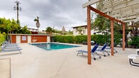 Located at 3902-3920 Lenawee Ave in Culver City, California, this outdoor amenity space centers around a generously sized swimming pool with clear, blue water and a classic tiled border. The pool deck is finished in light-toned concrete, providing ample space for relaxation and movement throughout the area. A row of blue lounge chairs lines one side of the pool, offering residents a comfortable place to unwind, while a wooden pergola structure at one end adds architectural interest and a shaded gathering spot. A prominent red-sided utility building sits at one end of the pool area, housing what appear to be restroom and storage facilities. The building is equipped with pool safety equipment, including a life ring and posted pool rules, reflecting a well-maintained and safety-conscious environment. The perimeter of the pool area is enclosed by a dense green hedge, providing privacy and a clean visual boundary. Tall palm trees rise above the surrounding landscape, lending a distinctly Southern California character to the space. The overall setting is open-air and single-level, designed to maximize usability and comfort. The combination of practical amenities, thoughtful landscaping, and a spacious pool deck makes this an inviting outdoor retreat in the heart of Culver City.