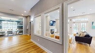 Located at 100 Town Walk Dr in Hamden, Connecticut, this thoughtfully designed single-floor clubhouse serves as a premier community amenity space. The layout flows seamlessly across several distinct areas, each finished with warm hardwood flooring, recessed lighting, and a cohesive contemporary aesthetic throughout.

The main lounge area features a classic white fireplace mantel with a wall-mounted flat-screen TV, complemented by comfortable seating arrangements on patterned area rugs. An adjacent seating room with elevated ceilings and a coffered tray ceiling detail offers a more intimate gathering space, furnished with a plush white sectional sofa and rich blue-gray walls.

A dedicated game room, painted in a vibrant teal with beadboard wainscoting, accommodates a full-size ping-pong table and receives ample natural light through large windows overlooking the outdoor pool area. The space connects visually to other areas through interior pass-through windows framed in crisp white trim.

The kitchenette is a standout feature, boasting bold periwinkle-blue cabinetry with gold hardware, quartz countertops, an undermount sink, and a kitchen island with bar seating — all illuminated by modern globe pendant lighting. Large windows and multiple sets of French doors open directly to the outdoor pool and patio, flooding the interior with natural light and providing effortless indoor-outdoor connectivity.