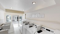 This single-story shared laundry facility is designed for practical, efficient use. The room is lined with multiple top-loading washing machines arranged in a row along one wall, providing ample capacity for residents. Alongside the machines, utility sinks offer additional convenience for hand-washing or pre-treating garments. Informational notices and posted guidelines are displayed on the wall above the machines, keeping users informed of proper usage and facility rules. The space is finished with light-colored ceramic tile flooring and cream-painted walls, contributing to a clean, bright atmosphere. Overhead flush-mounted light fixtures provide consistent illumination throughout. At the far end of the room, a gated entryway opens to an outdoor area, allowing for natural light and ventilation to flow into the space. The layout is straightforward and functional, prioritizing ease of movement and accessibility. Overall, the facility presents a well-maintained, no-frills environment suited to everyday laundry needs.
