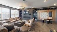 This single-story community space in Murfreesboro, Tennessee, is designed with a contemporary aesthetic that balances functionality and comfort. Large windows with horizontal blinds and curtain panels line the main gathering area, flooding the space with natural light and creating an open, welcoming atmosphere. The flooring throughout consists of large-format tile in a warm neutral tone, providing a cohesive and durable foundation across all areas.

The lounge area serves as a central focal point, anchored by a painted brick accent wall housing a mounted flat-screen television above a sleek linear fireplace. Built-in shelving with cross-hatch detailing flanks the fireplace, offering display space and built-in game features including a wall-mounted Scrabble board. An elegant chandelier overhead adds a refined touch to the seating arrangement, which includes plush chairs and a variety of accent furnishings.

Adjacent areas include a casual dining and high-top seating zone, ideal for social gatherings, as well as a dedicated game room featuring a foosball table. A reception counter with a dark countertop and pass-through window separates functional zones, while glass-paneled sliding doors lead to additional private or meeting spaces, completing this well-appointed community hub.