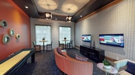 This well-appointed community lounge and recreation room is designed for both socializing and entertainment, offering a dynamic blend of bold design elements and practical amenities. The space is anchored by a striking terracotta-red accent wall adorned with a curated arrangement of circular decorative mirrors and a large framed art piece, lending the room a vibrant, gallery-like character. A full-length shuffleboard table runs alongside this feature wall, providing a central recreational focal point.

The entertainment area opposite features a textured houndstooth-patterned accent wall finished with a warm wood trim border, against which two flat-screen TVs are mounted side by side above a sleek black media console. Curved coral-upholstered seating and a neutral accent chair are arranged for comfortable viewing, complemented by patterned carpeting in deep navy and charcoal tones that flows throughout the space.

Natural light enters through multiple large gridded windows, each accompanied by high-top bistro tables and bar chairs — ideal for casual conversation or individual use. The tray ceiling is fitted with a dark contrasting inset and outfitted with geometric pendant light fixtures that contribute to the room's contemporary aesthetic. Glass double doors provide direct outdoor access, completing this thoughtfully designed, multi-use gathering space.