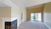 This single-floor residence offers a well-appointed living space defined by clean lines and a warm, neutral color palette. The living room serves as the heart of the home, featuring a classic tile-surround fireplace with a decorative metal screen that anchors the space with both style and function. Plush wall-to-wall carpeting adds comfort underfoot, while crown molding along the ceiling lends an elegant architectural touch throughout.

Natural light flows generously into the living area through a large sliding glass door, which opens onto a balcony framed by a metal railing. The outdoor view reveals verdant greenery and mature palm trees, creating a serene and tropical backdrop that enhances the overall ambiance of the interior.

The vaulted ceiling in the living area contributes to an airy, open feel, making the space feel expansive and welcoming. A hallway visible from the living room connects to additional areas of the home, suggesting a thoughtful and functional layout.

The overall aesthetic is light, clean, and livable — a harmonious blend of comfortable design elements and natural surroundings that makes this space both practical and inviting.