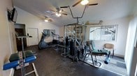 This dedicated fitness center offers a comprehensive workout environment designed to accommodate a wide range of exercise needs. The space is anchored by a large multi-station cable machine, providing users with numerous strength training options in a single unit. Complementing this centerpiece are multiple cardio machines, including an elliptical trainer and a treadmill, positioned to take advantage of the natural light filtering through the vertical-blind-covered windows. Free weights and additional training accessories are also available throughout the room. The flooring features durable rubber matting, ideal for high-impact activity and equipment stability. Two ceiling fans keep air circulating throughout the room, contributing to a comfortable workout environment. The textured ceiling and vaulted roofline lend a sense of openness to the space. A door on one wall provides convenient access, while the overall layout ensures sufficient room to move between stations. The combination of strength, cardio, and functional training equipment makes this a versatile and fully functional fitness facility suited for a variety of training routines.