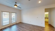 This single-floor unit presents a cohesive, modern design characterized by wood-look flooring that flows consistently throughout the space. The open-concept living and kitchen area forms the heart of the unit, where a well-appointed kitchen features flat-panel white cabinetry extending to the ceiling, dark countertops, a subtle hex-tile backsplash, and a full suite of stainless steel appliances including a refrigerator with ice dispenser, range, and built-in microwave. A kitchen island with pendant lighting provides additional counter space and defines the transition into the living area. The living area is generously proportioned, with a ceiling fan, recessed lighting, and a glass door that introduces natural light and outdoor views. Bedroom spaces are well-sized, each equipped with ceiling fans and wood-look flooring. The bathroom is cleanly designed with a walk-in glass-enclosed shower featuring white subway tile, a white vanity with dark countertop and matte black hardware, and a mirror with overhead lighting. A dedicated laundry room includes a stacked Whirlpool washer and dryer unit alongside built-in shelving, adding functional convenience. Throughout, bright white walls and clean lines reinforce the unit's contemporary character.