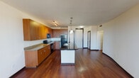 Located at 1101 Bear Creek Pkwy in Keller, Texas, this single-floor unit offers a cohesive, open-concept layout with consistent rich hardwood flooring flowing throughout the entire space. The kitchen serves as a central highlight, featuring warm medium-toned wood cabinetry extending to the ceiling for ample storage, granite-look countertops, and a suite of black appliances including a range, microwave, and refrigerator. A peninsula island with a dark countertop and pendant lighting above provides both additional workspace and a natural division between the kitchen and living areas. Track lighting illuminates the kitchen workspace effectively.

The living area is generously proportioned, with neutral walls, a ceiling fan, and a bank of windows that invite natural light and provide views of the surrounding area. The curved architectural wall detail adds a subtle design element to the otherwise clean-lined interior. The layout includes a hallway corridor leading to additional rooms, including a bathroom fitted with a single-sink vanity with white countertops, a large mirror with built-in shelving, and dark hardwood flooring continuing from the main living spaces. The overall ambiance is clean, modern, and functional, with a neutral palette that complements the warm wood tones throughout.