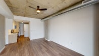 This single-floor unit showcases a compelling blend of industrial and contemporary design elements. The open living area immediately draws attention with its exposed concrete ceiling, visible cylindrical ductwork, and a ceiling fan that adds both function and character. Warm wood-look flooring runs throughout, complementing the raw concrete columns and crisp white walls to create a balanced, urban aesthetic. Natural light enters through a window, offering views of the surrounding cityscape.

The galley-style kitchen is efficiently laid out and well-appointed with stainless steel appliances, including a Whirlpool refrigerator, electric range, over-the-range microwave, and dishwasher. Cabinetry combines high-gloss white upper cabinets with light wood-toned lower units and tall pantry storage. A white subway tile backsplash, undermount sink with a matte black faucet, and light quartz countertops complete the modern aesthetic. Track lighting illuminates the workspace effectively.

The bathroom continues the cohesive design language, featuring a tub-shower combination with vertically stacked white tile surround, matte black fixtures and hardware, a light wood vanity with an undermount sink, white countertop, and a large mirror. In-unit laundry is also present, adding everyday convenience to the thoughtfully designed layout.