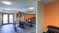 This single-floor fitness center is designed to provide a comprehensive workout environment for its users. The space is immediately distinguished by its bold orange accent walls, which add energy and vibrancy to the room, contrasting cleanly with the neutral white ceiling and light-colored walls on the opposite side. Overhead flush-mount lighting ensures the entire area is well-illuminated throughout.

The facility is outfitted with a multi-station cable and weight machine, offering a variety of strength training exercises from a single unit. Complementing the strength equipment, the cardio section features multiple treadmills equipped with individual digital screens, allowing users to track their workouts with ease.

The flooring consists of durable, dark rubberized material, which is standard for fitness spaces and provides both cushioning and grip during exercise. The layout is thoughtfully organized, with strength training equipment positioned toward the front and cardio machines arranged deeper into the space, creating a logical flow for users moving through their routines.

Overall, the fitness center presents a functional and motivating atmosphere, combining practical equipment placement with an energetic color scheme to create an inviting space for exercise.