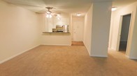 This single-floor unit presents an open-concept layout where the living area and kitchen are seamlessly connected, creating a cohesive and practical living environment. The living space features warm, neutral-toned carpet underfoot and smooth, light-painted walls that contribute to a bright and welcoming ambiance. A ceiling fan with an integrated light fixture is centrally positioned overhead, providing both air circulation and illumination throughout the space.

The kitchen is accessible directly from the living area via a raised breakfast bar counter finished in a dark laminate surface, which serves as both a functional workspace and a casual dining or seating area. The kitchen is equipped with stainless steel appliances, including a refrigerator and a range, and is complemented by white cabinetry that extends to the upper portion of the wall. The cabinetry provides ample storage, and the overall kitchen design maintains a clean, classic aesthetic.

Adjacent to the kitchen, a hallway leads to the unit's entry door, where hard flooring transitions from the carpeted living area. The entry area is well-lit with a flush-mount ceiling light, and the white-paneled door adds a clean, finished look to the space.