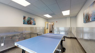 This single-floor recreational room is purpose-built for active use, centered around a full-size Cornilleau ping pong table finished in light blue. The room's most distinctive architectural detail is the diamond-plate stainless steel wainscoting that lines the lower half of all walls, providing a durable, industrial aesthetic that contrasts with the smooth, neutral-toned upper walls. Large-format tile flooring in a dark wood-look finish runs throughout, adding warmth to the otherwise contemporary interior. The suspended ceiling incorporates evenly spaced recessed LED panel lights, ensuring consistent, well-distributed illumination across the entire play area. A colorful abstract canvas painting serves as the primary wall decoration, introducing a creative element to the space. A window on one side allows natural light to supplement the artificial lighting. Access to the room is provided through a light wood-framed door with a frosted glass panel, which leads to an adjacent corridor. The room offers ample clearance on all sides of the table for unrestricted play. Overall, the space balances functional durability with a clean, modern design suitable for recreational or amenity use.