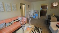 This single-floor residence offers a comfortable and functional layout with an open-concept design that connects the living area to the kitchen. The living room is highlighted by warm beige walls and carpeted flooring, creating a cozy atmosphere. A vibrant orange sofa anchors the seating area, complemented by a rectangular coffee table resting on a bold black-and-white polka dot area rug, adding a playful contrast to the neutral surroundings. Decorative tall branches with black-and-white accents serve as a striking focal point in the room. A low-profile cubby storage unit near the hallway provides practical organization with a pop of blue color. The space flows naturally toward a hallway leading to additional rooms, as well as toward the kitchen, which features dark cabinetry and counter space visible from the living area. Wall art adds personality to the neutral palette throughout. The overall ambiance is casual and inviting, balancing functional living with personalized decorative touches that give the space a lived-in, homey character.