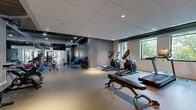 Located at 915 Washington Ave N in Minneapolis's vibrant North Loop neighborhood, this fitness center offers a comprehensive workout facility within a thoughtfully designed single-floor space. The gym features a modern aesthetic with a combination of light and dark design elements — deep charcoal accent walls contrast with lighter gray and neutral tones, while recessed lighting and exposed industrial ceiling details add character and depth throughout.

The space is organized to accommodate a wide range of exercise disciplines. A dedicated cardio zone lines the perimeter, housing treadmills, elliptical trainers, and cycling bikes, with windows overlooking greenery providing natural light and a refreshing outdoor connection. A free weights area includes a full dumbbell rack and benches, supported by wall mirrors that enhance the sense of openness. Blue exercise mats, stability balls, and kettlebells occupy a functional stretching and cross-training zone at the center of the floor.

Toward the entrance, a multi-functional cable machine serves as a focal point for strength training, flanked by a glass partition that visually connects to an adjoining corridor. A large wall-mounted flat-screen TV is centrally positioned for entertainment during workouts. Durable speckled rubber flooring spans the entire space, ensuring safety and comfort. Together, these features create a well-rounded, professionally appointed fitness environment.