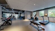 Located at 915 Washington Ave N in Minneapolis's vibrant North Loop neighborhood, this fitness center offers a comprehensive workout facility within a thoughtfully designed single-floor space. The gym features a modern aesthetic with a combination of light and dark design elements — deep charcoal accent walls contrast with lighter gray and neutral tones, while recessed lighting and exposed industrial ceiling details add character and depth throughout.

The space is organized to accommodate a wide range of exercise disciplines. A dedicated cardio zone lines the perimeter, housing treadmills, elliptical trainers, and cycling bikes, with windows overlooking greenery providing natural light and a refreshing outdoor connection. A free weights area includes a full dumbbell rack and benches, supported by wall mirrors that enhance the sense of openness. Blue exercise mats, stability balls, and kettlebells occupy a functional stretching and cross-training zone at the center of the floor.

Toward the entrance, a multi-functional cable machine serves as a focal point for strength training, flanked by a glass partition that visually connects to an adjoining corridor. A large wall-mounted flat-screen TV is centrally positioned for entertainment during workouts. Durable speckled rubber flooring spans the entire space, ensuring safety and comfort. Together, these features create a well-rounded, professionally appointed fitness environment.