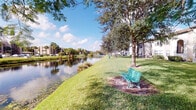 This single-story property is defined primarily by its picturesque outdoor surroundings. The exterior grounds front a calm, reflective waterway bordered by well-maintained green lawn, offering a serene and scenic environment. Mature trees, including native Florida species such as palms and live oaks, provide natural shade along the water's edge. A classic green bench positioned near the shoreline creates a dedicated outdoor relaxation spot with direct views across the lake. The landscaping is thoughtfully maintained, contributing to an overall atmosphere of quiet, natural tranquility. Neighboring low-rise buildings are visible in the distance across the water, suggesting a community-oriented setting. The surrounding environment is characteristic of a Florida waterfront community, with abundant greenery, open skies, and warm natural light enhancing the outdoor ambiance. Unfortunately, the interior images provided are too dark to allow for a detailed assessment of the indoor spaces, so a full interior description cannot be offered at this time. The combination of waterfront access, mature landscaping, and single-story accessibility makes this property well-suited for those who value outdoor amenity and a peaceful, nature-forward living environment.
