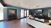 This single-story clubhouse offers a well-appointed communal gathering space designed for both relaxation and socializing. The entrance foyer greets visitors with glass French doors, warm wood-look flooring, a large decorative mirror, and a cozy seating area that sets an inviting tone throughout.

The main lounge area is generously proportioned, featuring a mix of teal, deep charcoal, and warm beige accent walls that create a modern yet comfortable atmosphere. A wall-mounted flat-screen TV, a sleek fireplace, and stylish white upholstered seating anchor the entertainment zone, while glass patio doors open to outdoor views and allow natural light to fill the space.

A sunlit dining nook, enclosed by floor-to-ceiling windows on multiple sides, provides panoramic views of the surrounding greenery and pool area. The space accommodates a round dining table with mid-century modern chairs beneath a statement pendant light.

Adjacent to the lounge, a kitchenette with wood-toned cabinetry, a breakfast bar with stools, and counter appliances offers convenient amenities for events and gatherings. Throughout, wood-look flooring, recessed lighting, and carefully selected artwork contribute to a cohesive, contemporary ambiance.