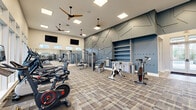 Located at 485 Crafton Cir in Charlottesville, VA, this expansive fitness center at Arch Brook Apartments spans a single floor and is divided into two distinct exercise rooms connected by a hallway, offering a comprehensive workout environment for residents. The main exercise room is bright and generously proportioned, featuring high ceilings, large windows along the perimeter that invite abundant natural light, and views of the surrounding community. A row of treadmills lines one wall, complemented by stationary bikes, ellipticals, and a full array of strength training machines. Floor-to-ceiling mirrors visually expand the space, while ceiling fans and recessed panel lighting ensure comfort. A bold geometric accent wall in steel blue adds a striking design focal point, alongside built-in shelving and a pegboard accessory station. The second exercise room offers a more intimate setting with light hardwood-style flooring, floor-to-ceiling mirrors, a spinning bike, a heavy punching bag, suspension trainers, and a full dumbbell rack. This room transitions seamlessly into a free-weight and functional training zone with rubber flooring, a rowing machine, a cable system, and a curved treadmill. Together, the two rooms provide a well-rounded, thoughtfully designed fitness facility suited for a wide range of training styles.