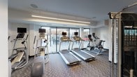 This fitness center offers a comprehensive workout environment within a modern, single-level layout. The space is defined by clean white walls, structural columns, and a bold chevron-patterned carpet that adds visual interest throughout. Linear LED lighting runs along the ceiling, casting bright, even illumination across the entire floor.

The cardio zone is positioned along the windowed wall, where a row of treadmills and elliptical machines equipped with individual monitors face floor-to-ceiling glass panels. These large windows overlook an outdoor terrace area, allowing natural light to fill the space and providing users with an open, airy feel during workouts.

The strength training section occupies the interior portion of the room, housing a variety of cable machines, cable crossover stations, and weight benches arranged to maximize usable floor space. Full-length mirrors are strategically placed to visually expand the area and assist with form. A dedicated storage unit with open wood shelving near the entry provides organized space for accessories or personal items.

A glass-paneled door leads directly to the adjacent outdoor terrace, enhancing connectivity between the indoor facility and exterior spaces. The overall design is functional, contemporary, and well-organized.