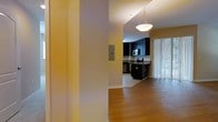 This single-floor residence offers a thoughtfully designed open layout that encourages a natural flow between living and functional spaces. The warm hardwood-style flooring runs throughout, lending a cohesive and inviting feel to the interior.

The kitchen is a standout feature, showcasing rich dark cabinetry that contrasts elegantly with the light-toned walls. Stainless steel appliances, including a microwave and range, are seamlessly integrated into the cabinetry, while a peninsula with dark countertops provides additional prep space and a casual dining or gathering area. A pendant light fixture overhead adds focused illumination and a touch of modern style.

Natural light is a key element of the space, with a large sliding glass door fitted with vertical blinds inviting abundant daylight into the kitchen and adjacent areas. The bright, neutral wall palette further enhances the sense of openness and airiness throughout.

A hallway connects the spaces with clean architectural lines, featuring a recessed utility panel flush to the wall, maintaining a tidy and uncluttered aesthetic. Overall, this residence combines functional design with warm, welcoming finishes to create a comfortable and practical living environment.