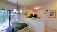 This single-floor residence offers a thoughtfully designed open-concept layout that seamlessly connects the living, dining, and kitchen areas. The living space is warm and inviting, filled with natural light that streams through large sliding glass doors, which provide direct access to a private outdoor patio surrounded by mature, verdant trees — creating a peaceful, nature-immersed backdrop.

The dining area sits adjacent to the living room and is anchored by a dark circular dining table paired with upholstered chairs featuring nailhead trim detailing. A elegant multi-arm chandelier with frosted glass shades hangs overhead, adding a refined touch to the space.

The kitchen is positioned just beyond the dining area and features a raised pass-through counter that maintains visual connectivity with the rest of the open floor plan. White upper cabinetry and black appliances, including a refrigerator and range, give the kitchen a clean, classic contrast. A small shelf along the pass-through provides additional display or prep space.

Throughout the main living areas, light-toned carpet and soft, neutral wall colors create a cohesive and calming ambiance. The overall design balances comfort and functionality, with abundant natural light and greenery views enhancing the sense of openness throughout the home.