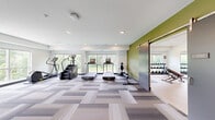 Located at 7689 Hardwood Ave S in Cottage Grove, MN, this fitness facility is thoughtfully designed to support a wide range of workout routines across two exercise rooms. The main exercise area features patterned, multi-tone carpet tiles that add a modern aesthetic to the spacious floor plan. Large windows along the perimeter flood the room with natural light and provide views of the surrounding greenery, creating an energizing atmosphere for workouts. This primary space is outfitted with an extensive selection of equipment, including treadmills, a stair climber, a rowing machine, a cable cross machine, a power rack with a bench press, and a multi-station cable machine. A bold olive green accent wall adds a vibrant pop of color, complementing the clean white walls and recessed lighting overhead. A sliding barn-style door with frosted glass panels connects the main area to a dedicated studio room. This secondary space features smooth hardwood flooring and a full-width wall mirror with a mounted flat-screen television, making it ideal for stretching, yoga, or guided workouts. The studio is further equipped with a dumbbell rack, medicine balls, a TRX suspension system, and a stationary bike. A water fountain and restroom access are also conveniently available within the facility.
