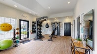 Located at 5744 Thorton St in Columbus, Ohio, this fitness center is a thoughtfully designed, single-story space built to accommodate a comprehensive range of workout routines. The interior features vaulted ceilings with recessed lighting, contributing to an open and energizing atmosphere throughout. Neutral-toned carpet flooring runs the length of the space, providing comfort underfoot while maintaining a clean, modern aesthetic.

The cardio zone is well-appointed with multiple treadmills, stationary bikes, and an elliptical trainer, each equipped with individual monitors for an engaging workout experience. Wall-mounted televisions are positioned throughout the room for added entertainment. Large windows with white plantation shutters line the walls, allowing abundant natural light to fill the space and offering views of the surrounding outdoor area, including a glimpse of a pool nearby.

The strength training area features a multi-functional cable machine, an adjustable weight bench, a speed bag, and a fully stocked dumbbell rack. A bold motivational word-art mural covers one wall, reinforcing an atmosphere of focus and determination. Floor-to-ceiling mirrors further enhance the sense of space. Stability balls and additional accessories round out the equipment offerings, making this fitness center a versatile and fully functional training facility.